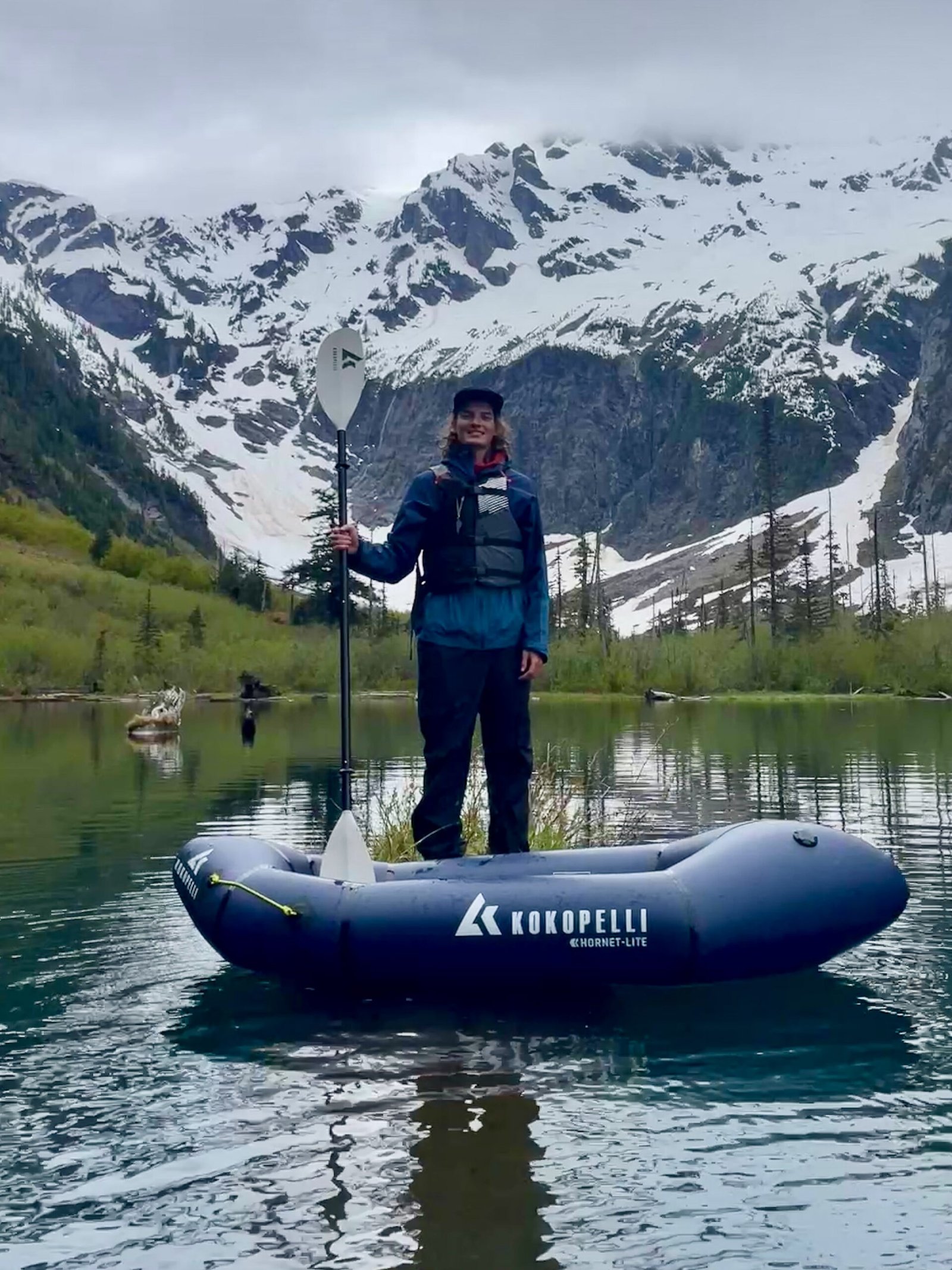 Packrafting guide Isaac Jackson standing in Goat Lake holding a paddle with North Cadet Peak in the background during an alpine lake adventure in Washington.