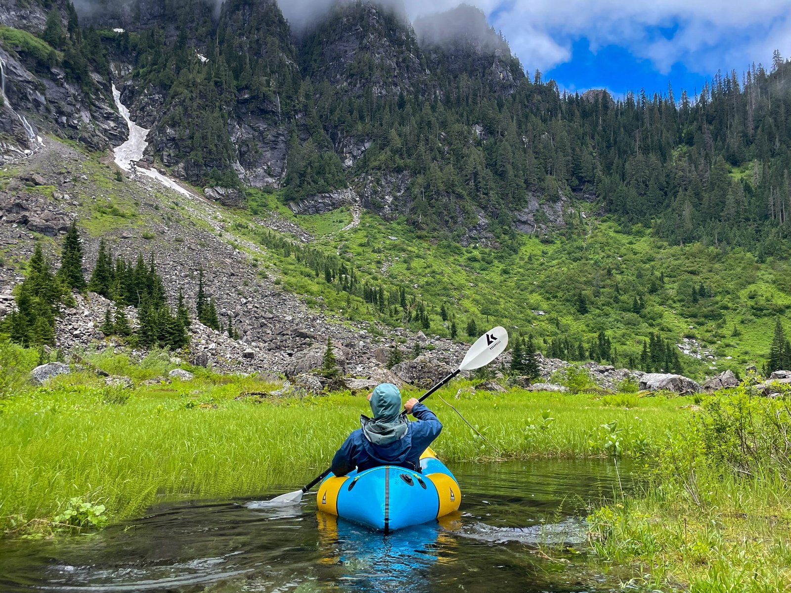 A guest paddling a packraft through an alpine stream with Cascade mountains in the background during the Optimum Adventures Lake 22 tour in Washington.