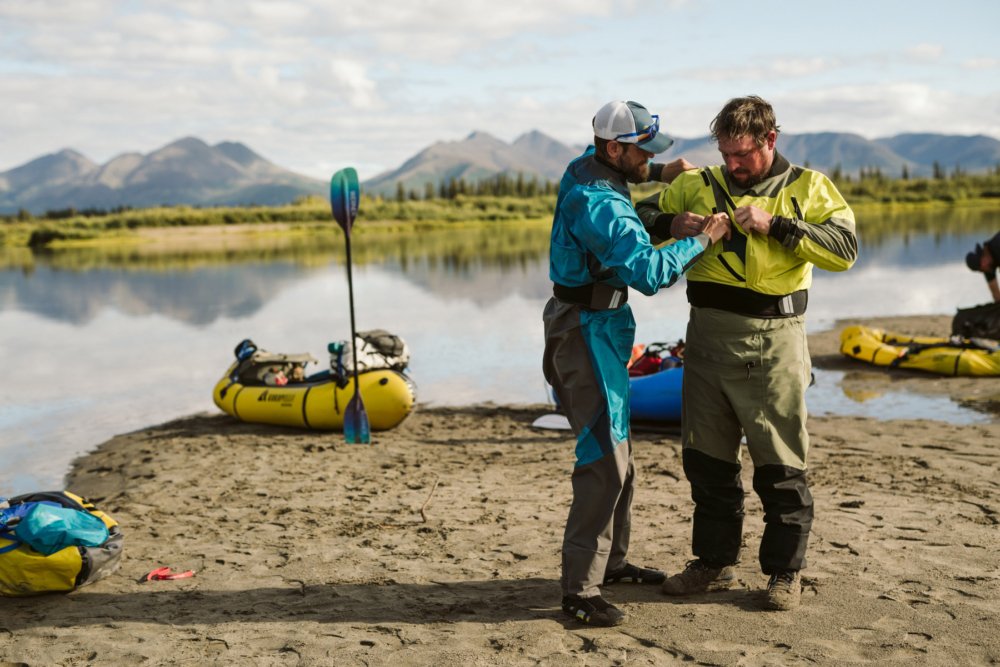 Two packrafters helping each other put on dry suits before a packrafting expedition, demonstrating dry suit rental options for cold-water recreation near Seattle.