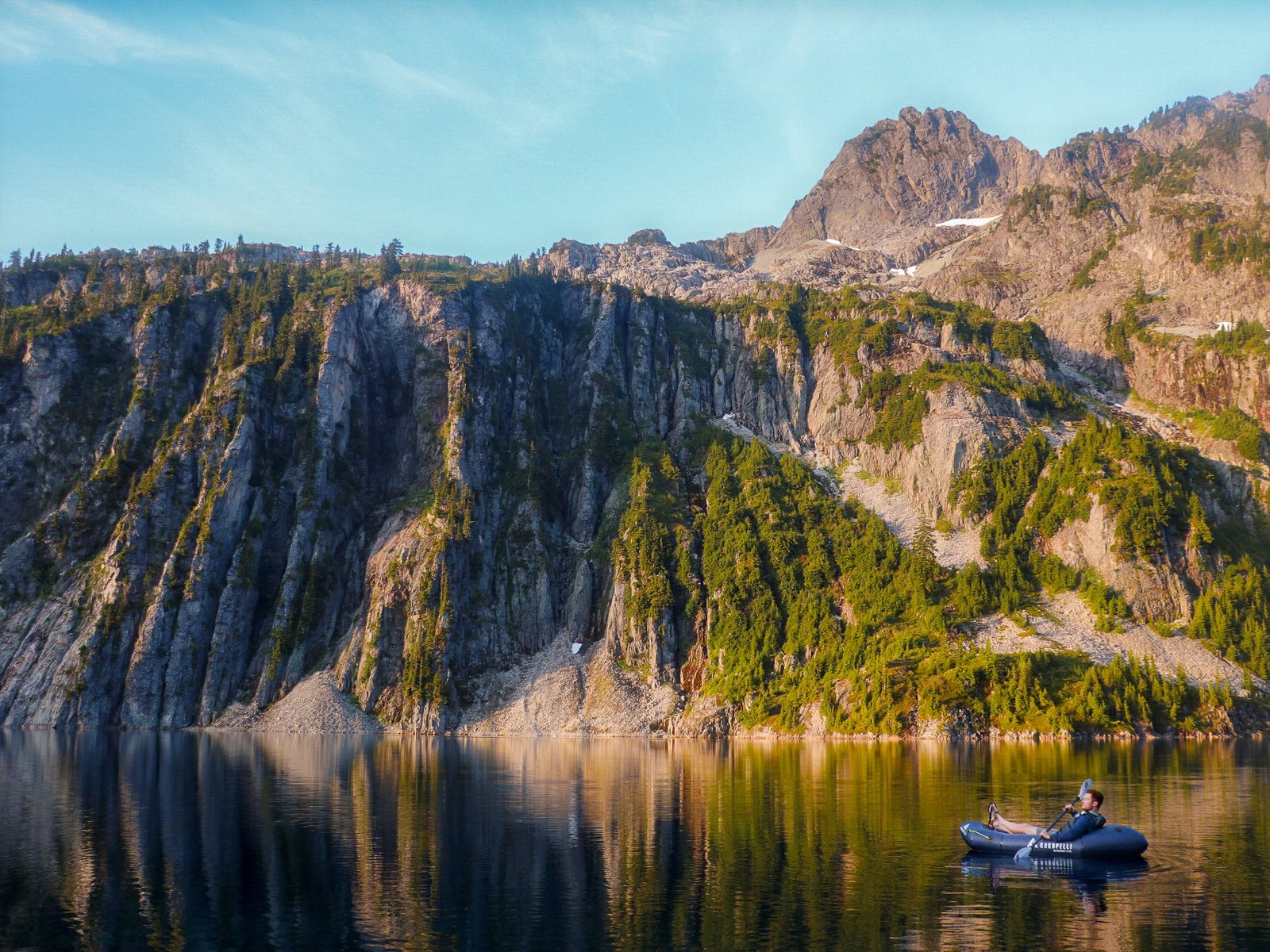Adam Jones, lead guide at Optimum Adventures, paddling a Kokopelli Hornet-Lite packraft on Snow Lake beneath blue skies and towering cliffs during the Snow Lake tour in Washington.