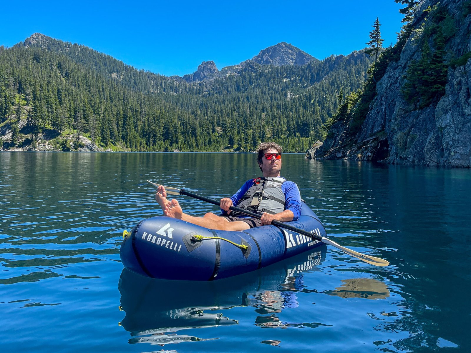A guest relaxing in a Kokopelli Hornet-Lite packraft on Snow Lake surrounded by evergreen forest, crystal-clear water, and mountain scenery during the Snow Lake kayaking tour.