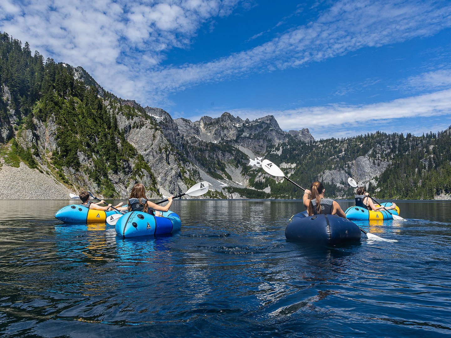 Guests paddling Alpacka and Kokopelli packrafts across Snow Lake during the Optimum Adventures Snow Lake tour with blue water, Cascade Mountains, and clear skies in Washington.