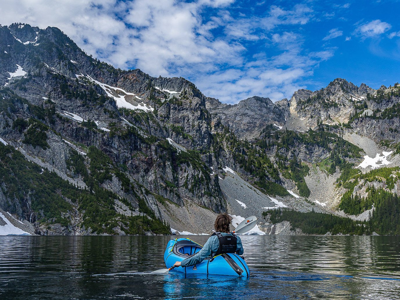A guest paddling an Alpacka Caribou packraft on Snow Lake with waterfalls, towering cliffs, and alpine peaks during the Optimum Adventures Snow Lake kayaking tour.